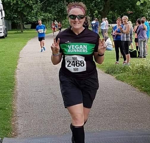 Ruth in her Vegan Runners top, crossing a race timing mat and smiling making a V symbol with both hands