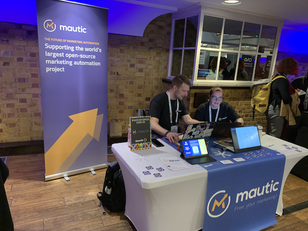 The Mautic stand at State of Open Con, with a rollup banner to the rear left, a white covered table with a blue roller which has the Mautic logo on it, and a screen showing a video of Mautic. The tablet also shows a landing page with a QR code. Two people are sitting behind the stand.