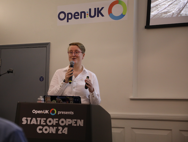 Photo of Ruth wearing a white, long sleeved shirt with the Mautic logo embroidered on it, holding a microphone and speaking at Open UK.